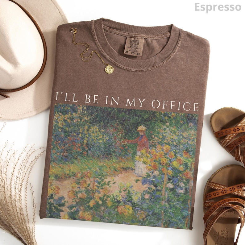 May include: Espresso-colored t-shirt with the text "I'll Be In My Office" above a colorful garden scene. A gold necklace and a hat are next to the shirt. Sandals are on the right side of the shirt.