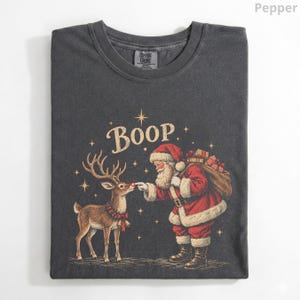 May include: A charcoal grey t-shirt featuring a graphic of Santa Claus touching the nose of a reindeer. The text Boop is printed above them, surrounded by small stars. Santa wears a red suit with white trim and carries a sack of gifts.