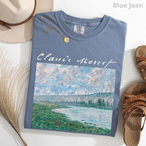 May include: A blue jean t-shirt with a white print of a painting by Claude Monet. The painting depicts a river scene with a cloudy sky and green trees.