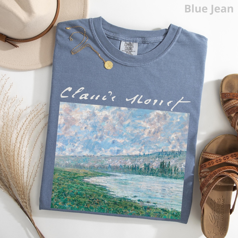 May include: A blue jean t-shirt with a white print of a painting by Claude Monet. The painting depicts a river scene with a cloudy sky and green trees.