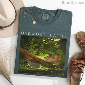 May include: Blue spruce-colored t-shirt with the text "ONE MORE CHAPTER" above a graphic of a person reading in a hammock. A gold necklace, a beige hat, and brown sandals are also in the image.