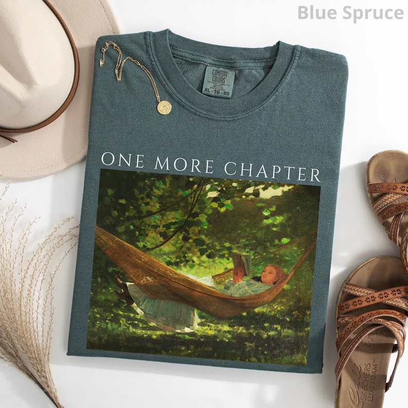 May include: Blue spruce-colored t-shirt with the text "ONE MORE CHAPTER" above a graphic of a person reading in a hammock. A gold necklace, a beige hat, and brown sandals are also in the image.