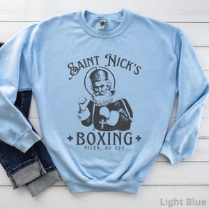 Op de afbeelding: Lichtblauwe trui met een afbeelding van Sint Nicolaas die boksen. De tekst luidt "Saint Nick's Boxing Nicaea, AD 325".
