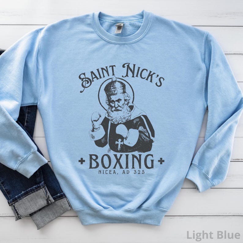 Op de afbeelding: Lichtblauwe trui met een afbeelding van Sint Nicolaas die boksen. De tekst luidt "Saint Nick's Boxing Nicaea, AD 325".