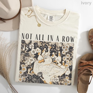 May include: Ivory-colored sweatshirt with the text "NOT ALL IN A ROW" above a graphic of ducks and flowers. A gold necklace and a beige hat are visible. Brown sandals are on the right side of the image.