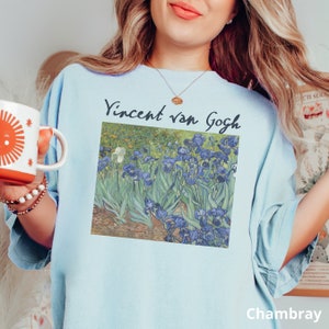 May include: A light blue t-shirt featuring a printed reproduction of Vincent van Gogh's Irises painting. The graphic displays a field of blue and purple irises with green stems and leaves, with the text Vincent van Gogh printed above the artwork.