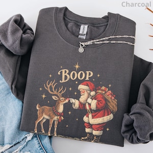 May include: A charcoal grey crewneck sweatshirt featuring a graphic of Santa Claus touching the nose of a reindeer. The text "Boop" is printed above the figures in a gold font, surrounded by small gold stars. A silver chain necklace rests on the collar.