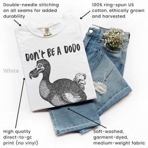May include: White t-shirt with a black graphic of a dodo bird and the text "DON'T BE A DODO." The shirt is paired with blue jeans. The shirt is made of 100% ring-spun US cotton and has double-needle stitching.