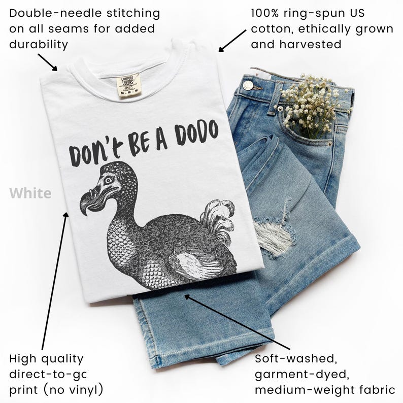 May include: White t-shirt with a black graphic of a dodo bird and the text "DON'T BE A DODO." The shirt is paired with blue jeans. The shirt is made of 100% ring-spun US cotton and has double-needle stitching.
