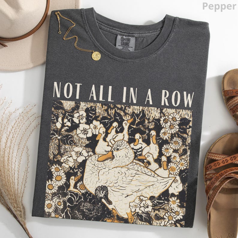 May include: A dark gray Comfort Colors t-shirt with the text "NOT ALL IN A ROW" above a graphic of ducks and flowers. A gold necklace and brown sandals are next to the shirt. The shirt is pepper colored.