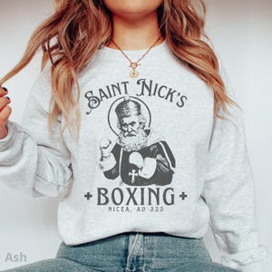 Op de afbeelding: Een grijs sweatshirt met een afbeelding van een cartoon Sinterklaas die boksen. De tekst luidt "Saint Nick's + Boxing + Nicaea, AD 325".