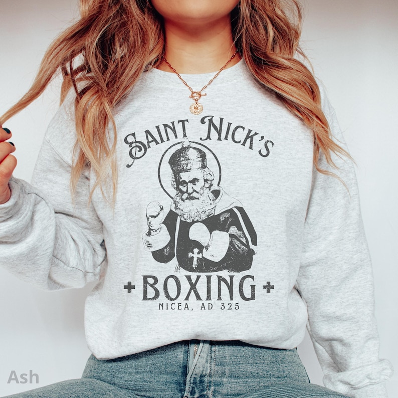 Op de afbeelding: Een grijs sweatshirt met een afbeelding van een cartoon Sinterklaas die boksen. De tekst luidt "Saint Nick's + Boxing + Nicaea, AD 325".