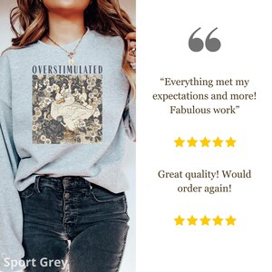 May include: Sport Grey sweatshirt with the word "OVERSTIMULATED" above a graphic of a white goose and other geese in a floral design. The sweatshirt is shown on a person.