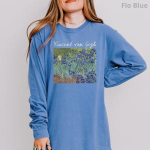 May include: A long-sleeve crewneck sweatshirt in a solid Flo Blue colour. The front features a rectangular graphic print of Vincent van Gogh's Irises painting, with the text "Vincent van Gogh" printed in a light yellow script font above the artwork.