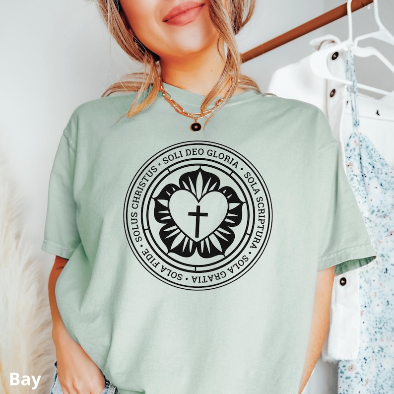 Puede incluir: Una camiseta verde claro con un dise&ntilde;o circular negro que presenta un coraz&oacute;n con una cruz en su interior. El c&iacute;rculo tiene el texto "SOLI DEO GLORIA - SOLA SCRIPTURA - SOLA GRATIA - SOLA FIDE - SOLUS CHRISTUS" alrededor del coraz&oacute;n.