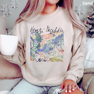 Puede incluir: Sudadera color arena con un estampado colorido de una pintura de paisaje de Henri Matisse. La obra de arte incluye tonos de verde, azul, amarillo y rojo. El nombre del artista est&aacute; impreso en cursiva negra encima de la obra.