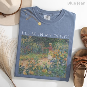 May include: Blue jean Comfort Colors t-shirt with the text "I'll Be In My Office" above a painting of a garden scene. A gold necklace and a beige hat are next to the shirt. Brown sandals are on the right.