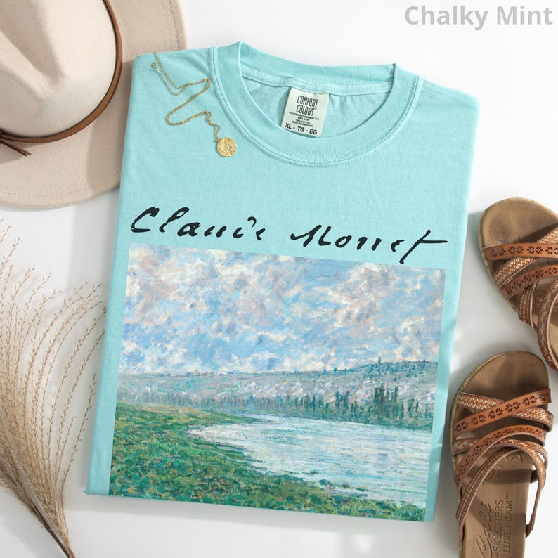 May include: A light blue t-shirt with a painting of a river and trees. The text "Classic Monet" is printed on the shirt in black.