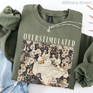 May include: Military green sweatshirt with the word "OVERSTIMULATED" printed in white. The graphic features ducks and flowers. A silver necklace with a sunburst pendant is on the sweatshirt. The sweatshirt is folded on top of a pair of blue jeans.