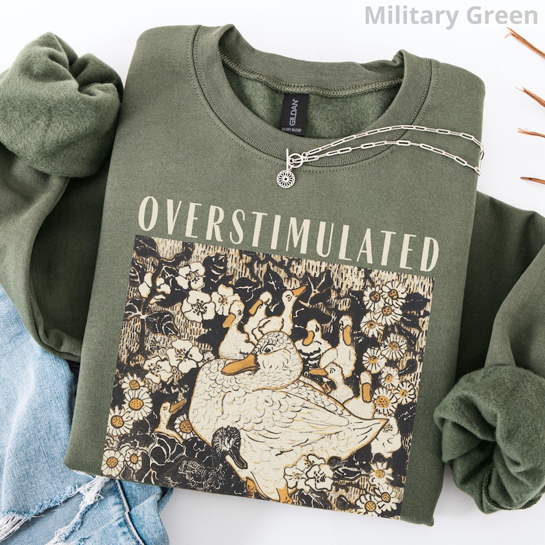May include: Military green sweatshirt with the word "OVERSTIMULATED" printed in white. The graphic features ducks and flowers. A silver necklace with a sunburst pendant is on the sweatshirt. The sweatshirt is folded on top of a pair of blue jeans.