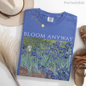 May include: A periwinkle blue t-shirt with the words "BLOOM ANYWAY" printed on the front. The shirt features a floral design with blue irises. A gold necklace and a beige hat are next to the shirt. Brown sandals are on the right.