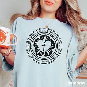 Puede incluir: Una camiseta azul claro con un gr&aacute;fico en blanco y negro de un coraz&oacute;n con una cruz dentro de un c&iacute;rculo. El c&iacute;rculo tiene el texto "SOLUS CHRISTUS SOLI DEO GLORIA SOLA SCRIPTURA SOLA GRATIA SOLA FIDE" alrededor.