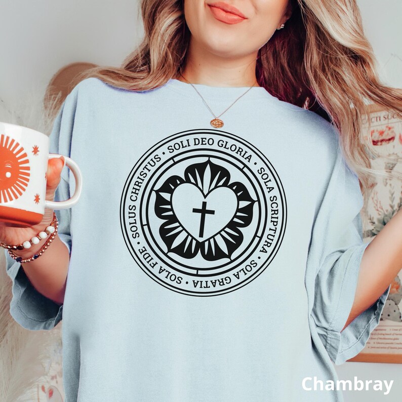 Puede incluir: Una camiseta azul claro con un gr&aacute;fico en blanco y negro de un coraz&oacute;n con una cruz dentro de un c&iacute;rculo. El c&iacute;rculo tiene el texto "SOLUS CHRISTUS SOLI DEO GLORIA SOLA SCRIPTURA SOLA GRATIA SOLA FIDE" alrededor.