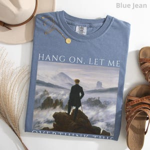 May include: A blue jean t-shirt with a white graphic of a painting. The painting depicts a man standing on a rock overlooking a sea of clouds. The text "Hang on, let me overthink this" is printed on the shirt.