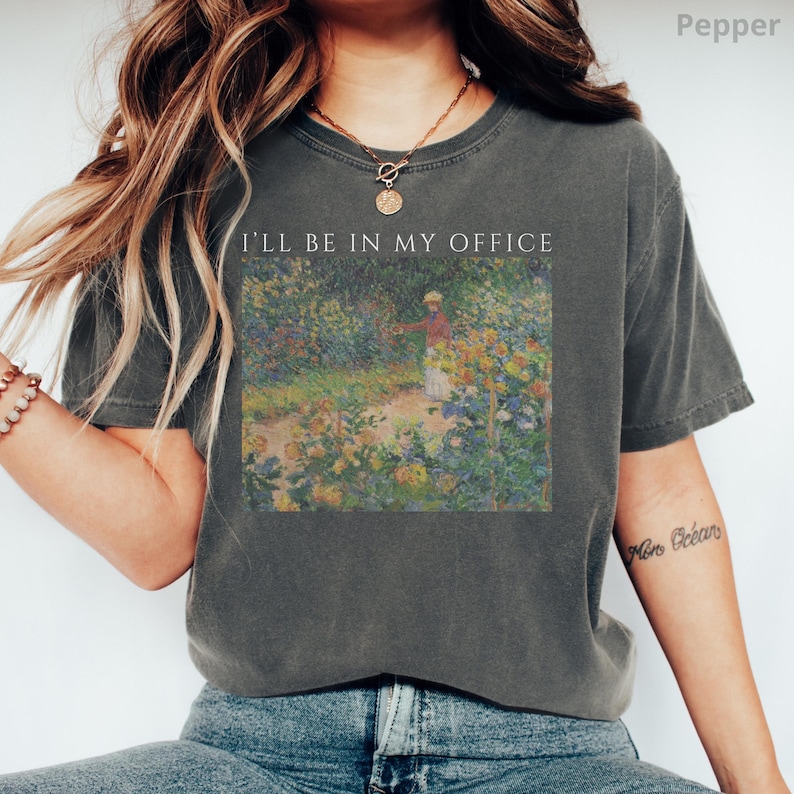 May include: Dark grey t-shirt with the text "I'LL BE IN MY OFFICE" above a colorful impressionist painting of a garden scene. The painting depicts a person in a garden surrounded by flowers.