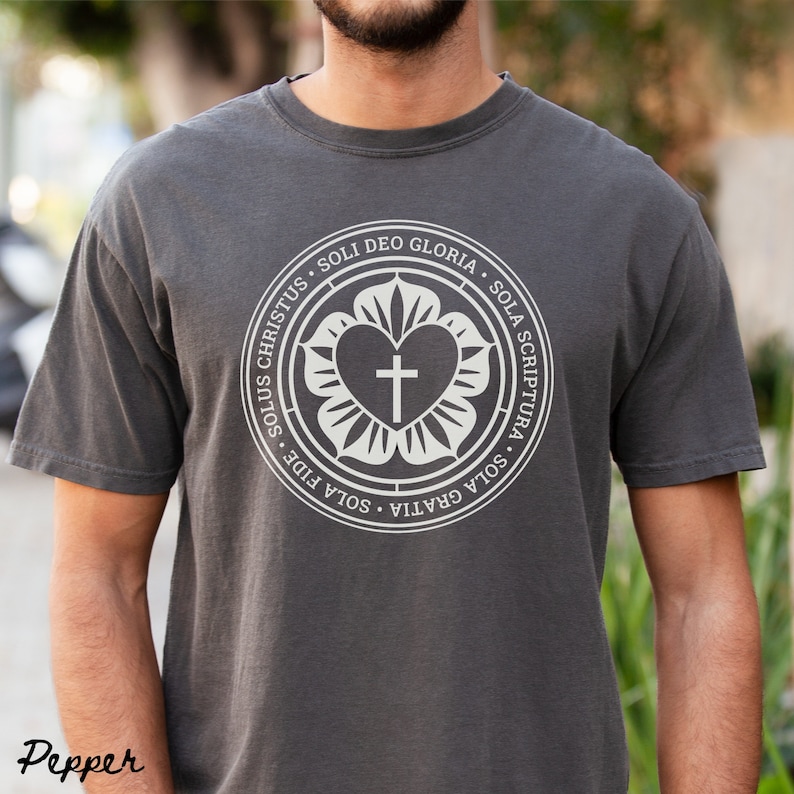 Puede incluir: Una camiseta gris antracita con un dise&ntilde;o gr&aacute;fico blanco. El dise&ntilde;o presenta un coraz&oacute;n con una cruz en su interior, rodeado de un c&iacute;rculo con el texto "Sola Gratia, Sola Fide, Solus Christus, Soli Deo Gloria, Sola Scriptura".