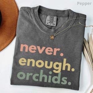 May include: A charcoal gray short-sleeve t-shirt with a crew neckline and a Comfort Colors brand label. The shirt features the text "never. enough. orchids." printed in three horizontal lines with coral, yellow, and sage green lettering.