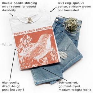 May include: White t-shirt with the text "RESPECT GOD'S CREATURES" and an orange bird graphic. The shirt is paired with light blue denim jeans. The t-shirt is made of 100% ring-spun cotton and has double-needle stitching.