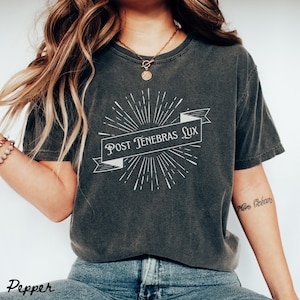 May include: A dark gray t-shirt with a white graphic design featuring a banner with the text "Post Tenebras Lux" and a sunburst design.
