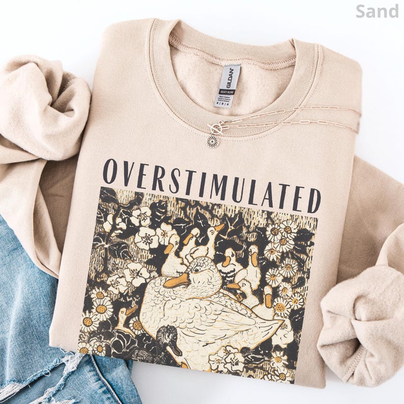 May include: Sand-colored sweatshirt with the word "OVERSTIMULATED" printed across the front. The graphic features a detailed illustration of ducks and flowers. A delicate silver necklace with a sun pendant is visible.