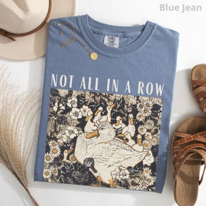 May include: Blue jean colored t-shirt with the text "NOT ALL IN A ROW". The graphic features white ducks and flowers. A gold necklace, a hat, and sandals are also visible.