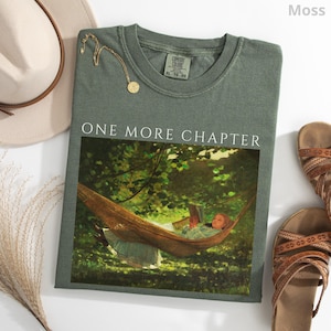 May include: A moss green t-shirt with the text "ONE MORE CHAPTER" above a painting of a person reading in a hammock. A gold necklace and a beige hat are next to the shirt. Brown sandals are on the right.