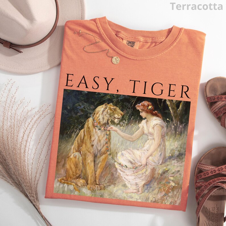 May include: Terracotta-colored t-shirt with the text "EASY, TIGER" above a painting of a woman and a tiger. A gold necklace and a hat are visible. Sandals are on the right side of the image.