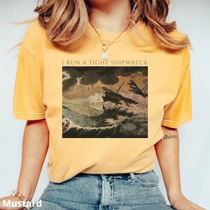 May include: Mustard yellow t-shirt with the text "I RUN A TIGHT SHIPWRECK" above a vintage illustration of a ship in a storm. The shirt is a solid color and has a relaxed fit.