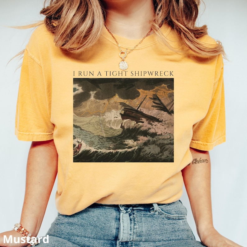 May include: Mustard yellow t-shirt with the text "I RUN A TIGHT SHIPWRECK" above a vintage illustration of a ship in a storm. The shirt is a solid color and has a relaxed fit.