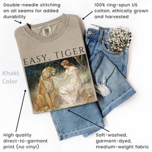 May include: Khaki-colored sweatshirt with the text "EASY, TIGER" and a graphic of a woman and a tiger. The sweatshirt is paired with blue jeans. Features include double-needle stitching, direct-to-garment print, and soft, medium-weight fabric.