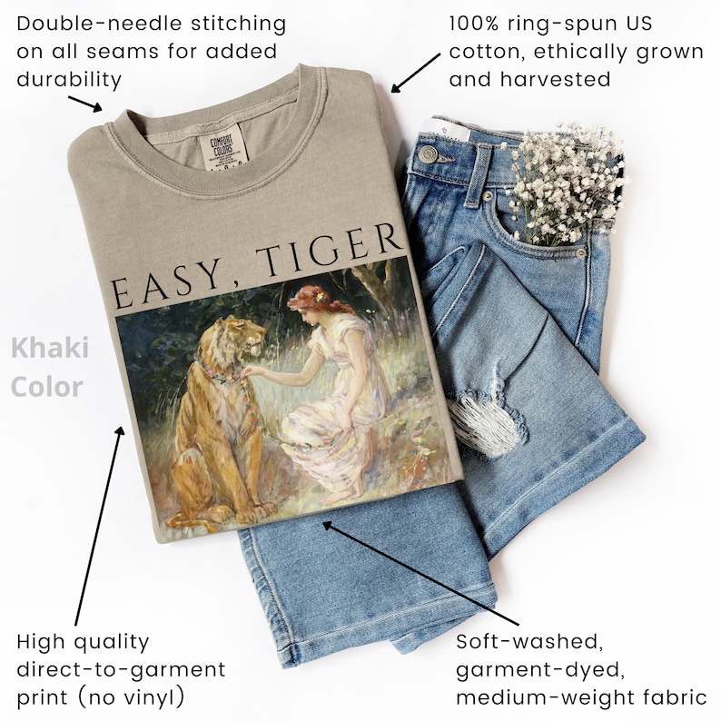 May include: Khaki-colored sweatshirt with the text "EASY, TIGER" and a graphic of a woman and a tiger. The sweatshirt is paired with blue jeans. Features include double-needle stitching, direct-to-garment print, and soft, medium-weight fabric.