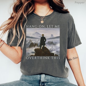 May include: A dark gray t-shirt with a graphic of a painting of a man standing on a mountaintop above the clouds. The text "Hang on, let me overthink this" is printed on the shirt.