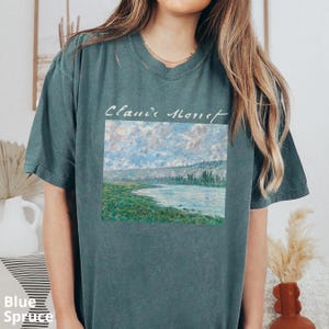May include: A dark teal t-shirt with a painting of a river and mountains. The text "Claude Monet" is printed above the painting.