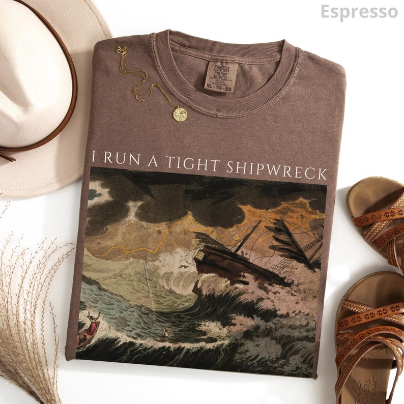 May include: Espresso-colored t-shirt with the text "I RUN A TIGHT SHIPWRECK" above a vintage illustration of a ship in a storm. A gold necklace and brown sandals are also visible.