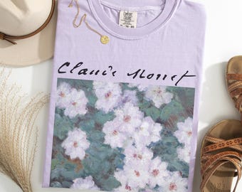 May include: A light purple t-shirt with a floral print of white flowers and the text "Claude Monet". A gold necklace and a beige hat are next to the shirt. The shirt is labeled "Orchid".