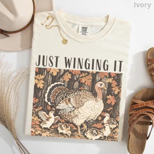 May include: An ivory t-shirt featuring the text JUST WINGING IT above a graphic of a turkey and several chicks in a forest with autumn leaves. The shirt has a Comfort Colors label at the neck and is displayed with a hat and sandals.