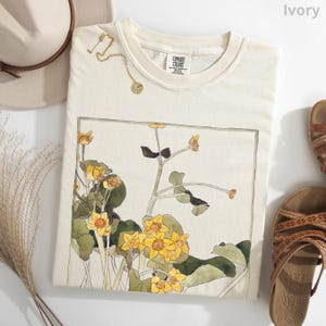 May include: An ivory t-shirt featuring a rectangular graphic print of yellow wildflowers with green leaves and stems. The shirt has a crew neckline with a Comfort Colors label. A gold-toned necklace with a circular pendant rests on the fabric.