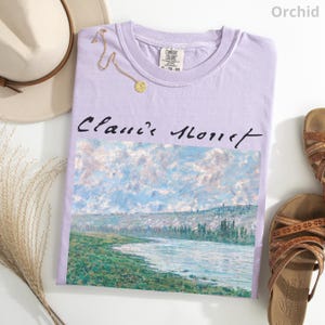 May include: A lavender t-shirt with a painting of a river and hills by Claude Monet. The text "Claude Monet" is printed in black on the shirt.