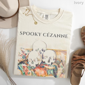May include: An ivory t-shirt featuring the text SPOOKY CÉZANNE in dark gray lettering above a graphic print of three skulls resting on a colorful, painterly fabric surface. The shirt has a crew neckline and a Comfort Colors brand label.