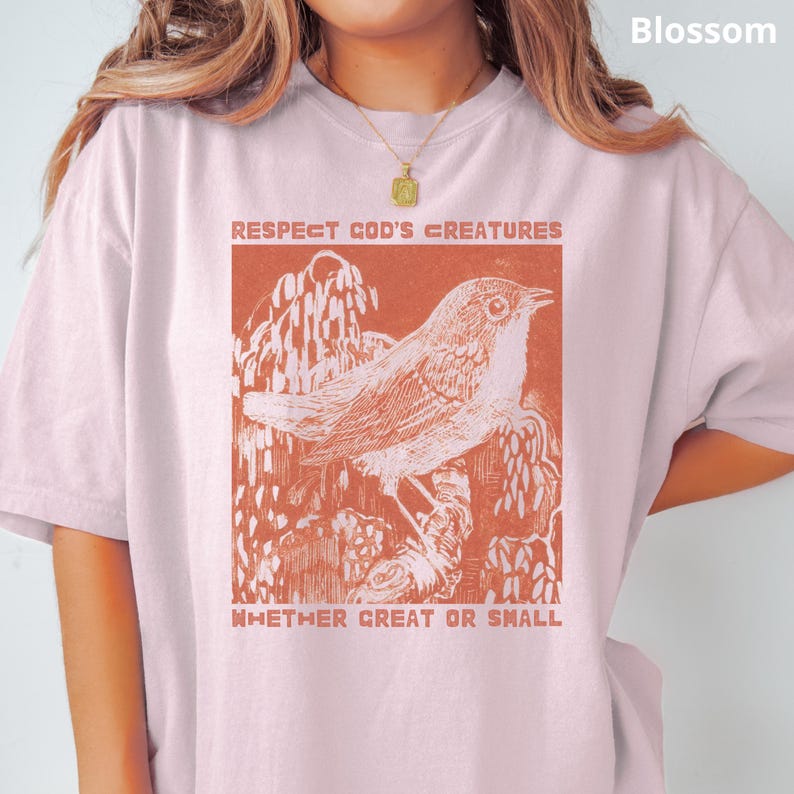 May include: Pale pink t-shirt with a rust-coloured illustration of a bird on a branch, and the text "RESPECT GOD'S CREATURES WHETHER GREAT OR SMALL". The shirt has short sleeves and a loose fit.